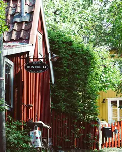 Röd trästuga med husnummerskylt omgiven av grönska