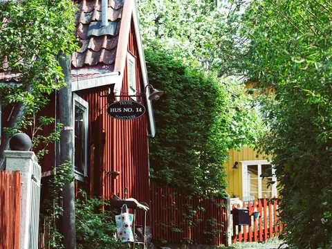 Röd trästuga med husnummerskylt omgiven av grönska
